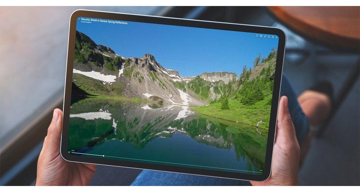 Person holding a tablet displaying a landscape scene of mountains, snow patches, green trees, and a reflective lake under a blue sky.