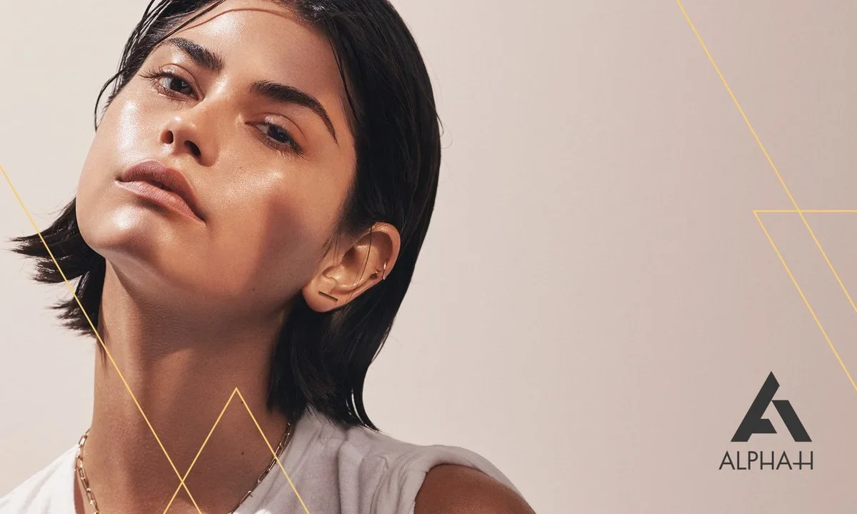 A woman with short dark hair, wearing a light-colored top, looking confidently at the camera against a neutral background with gold geometric lines and the logo 'ALPHA-H' in the bottom right corner.