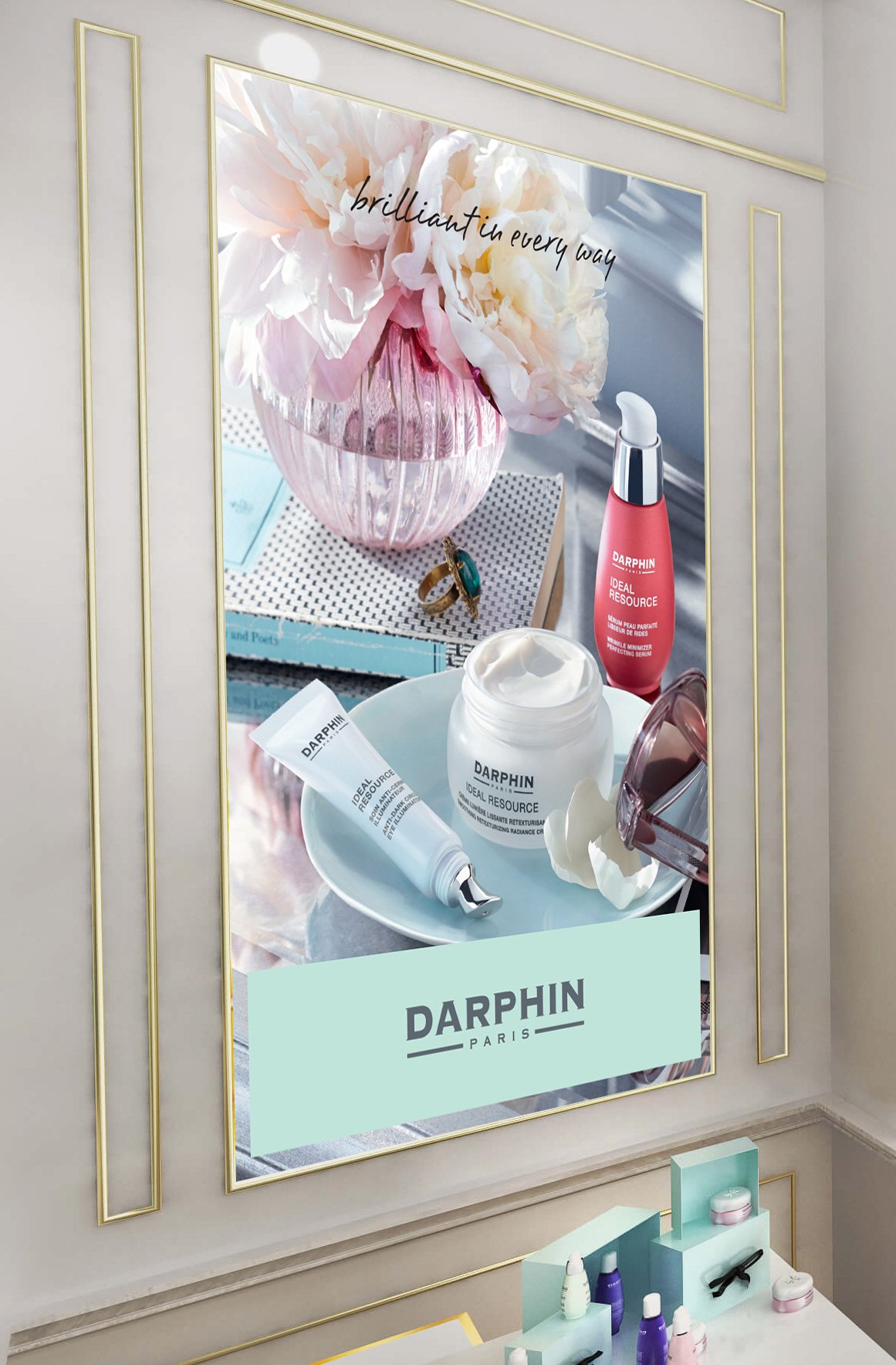 A large mirror with gold trim reflecting a display of skincare products, a pink vase with peonies, and a stack of books with jewelry. The mirror has the phrase 'brilliant in every way' written on it, and the display includes Darphin skincare items an